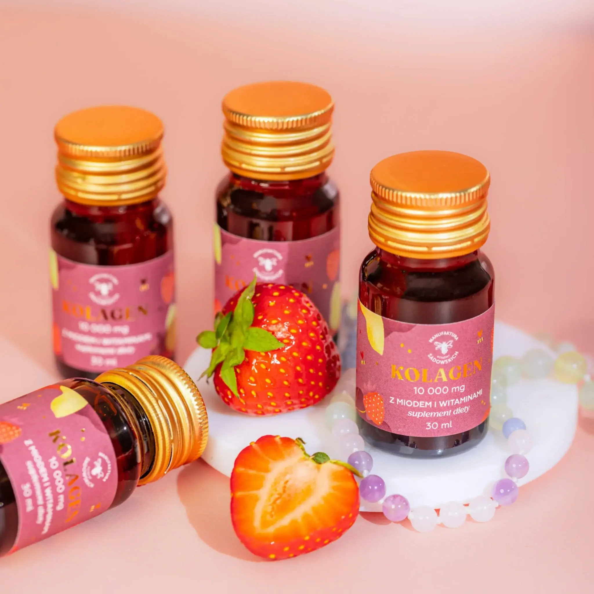 Zbliżenie na buteleczki kolagenu Manufaktura Sadowskich z miodem i witaminami, udekorowane truskawkami i koralikami na różowym tle.