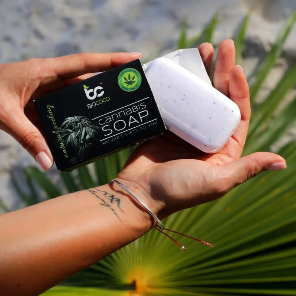 Mydło konopne BjoCoco z naturalnym peelingiem, trzymane w dłoniach na tle liścia palmowego, w naturalnym otoczeniu.