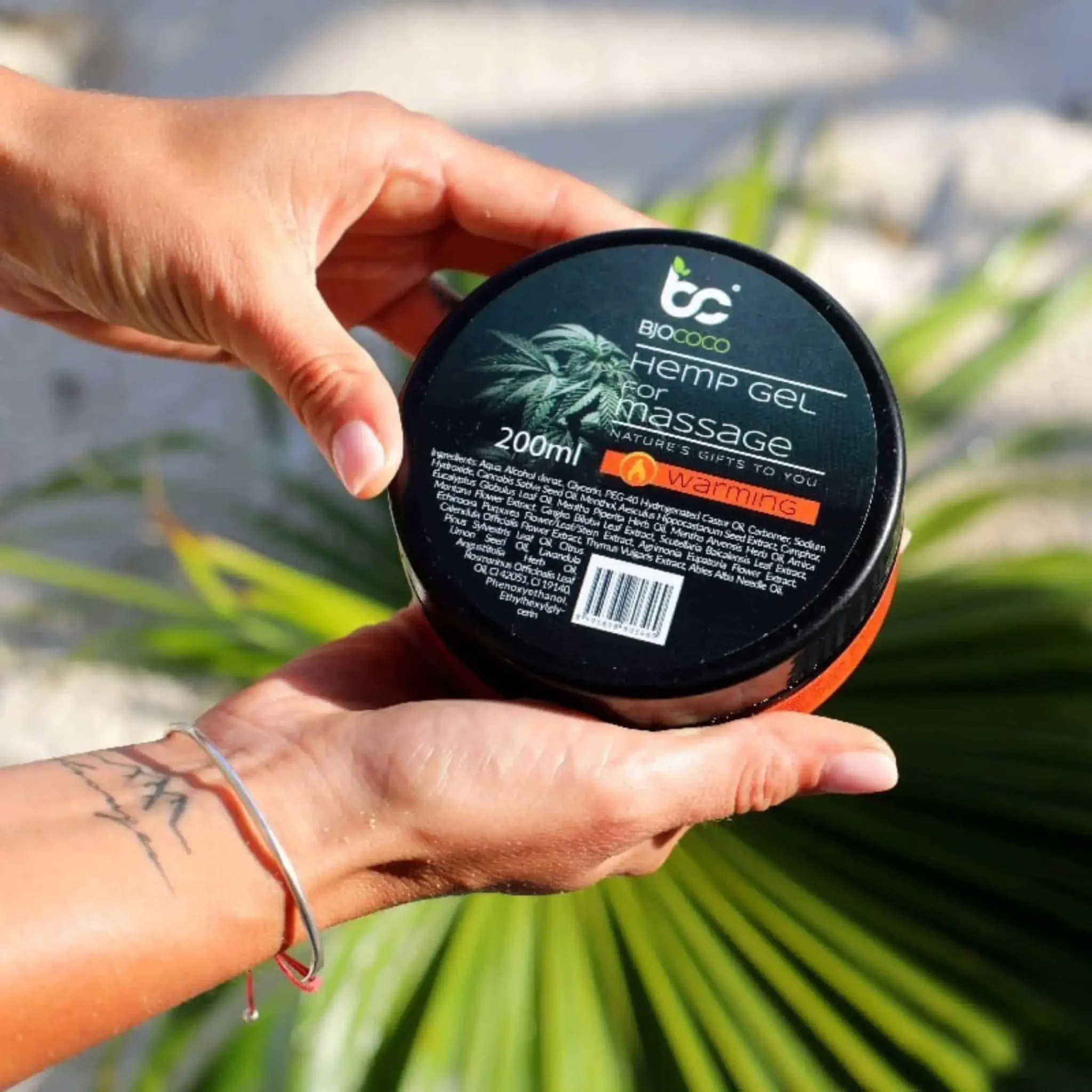 Opakowanie rozgrzewającego żelu konopnego BjoCoco o pojemności 200 ml, trzymane w kobiecych dłoniach z palmowymi liśćmi w tle.