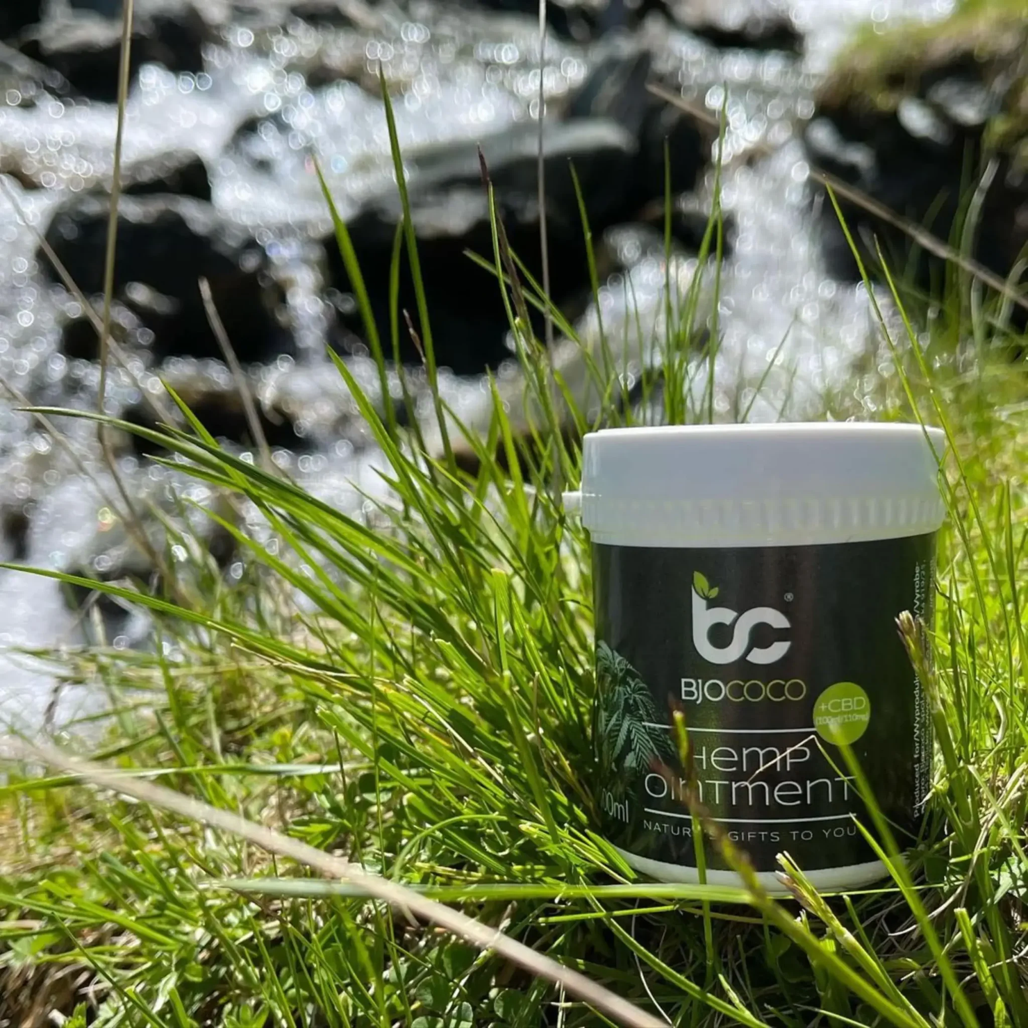 Maść konopna BjoCoco ustawiona w trawie na tle strumienia. Produkt z naturalnym CBD.