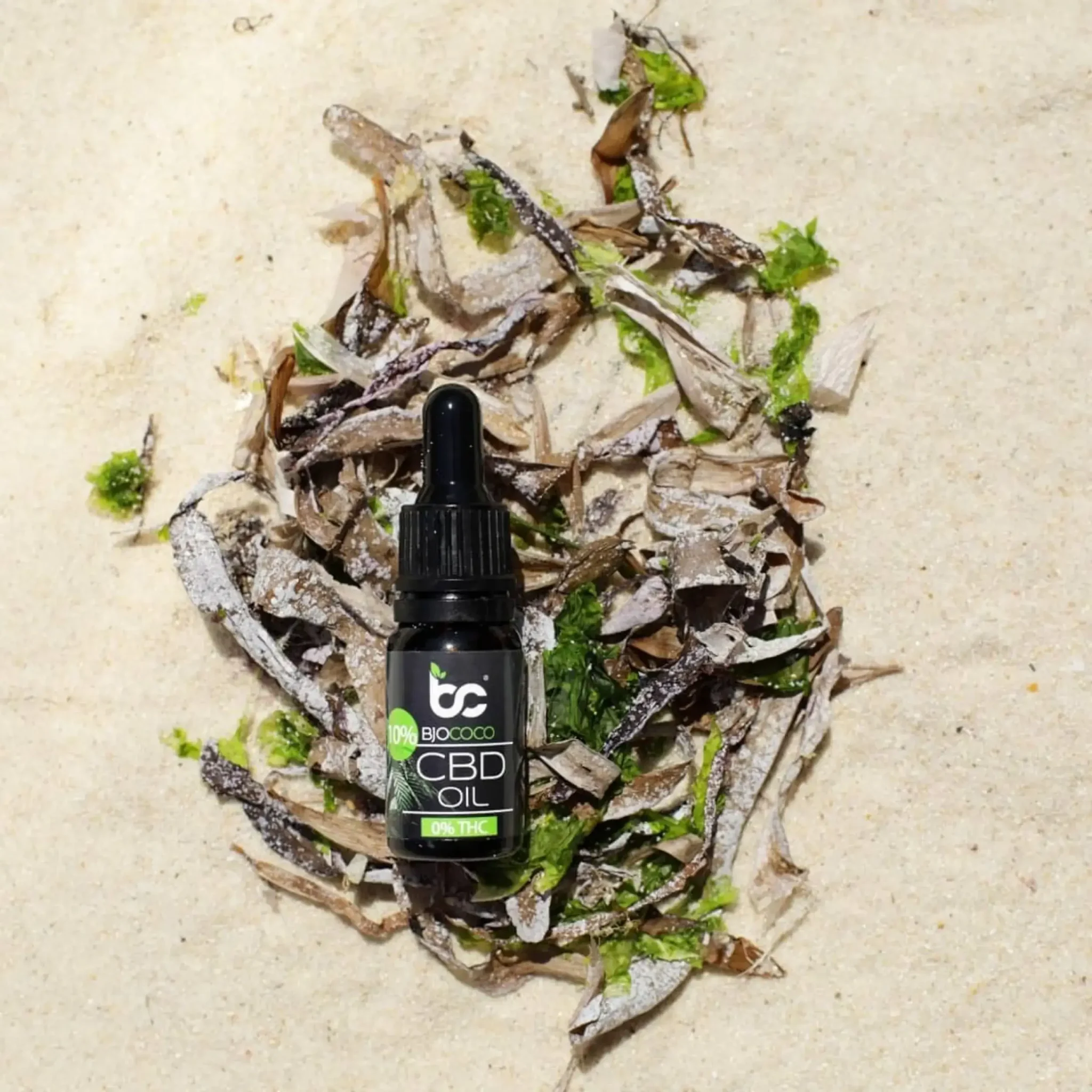 Olejek CBD marki BjoCoco umieszczony na piasku w otoczeniu wodorostów, podkreślający naturalność i organiczne pochodzenie produktu.